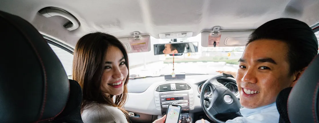Een jonge vrouw aan het stuur en een jonge man naast haar kijken lachend om vanuit de auto