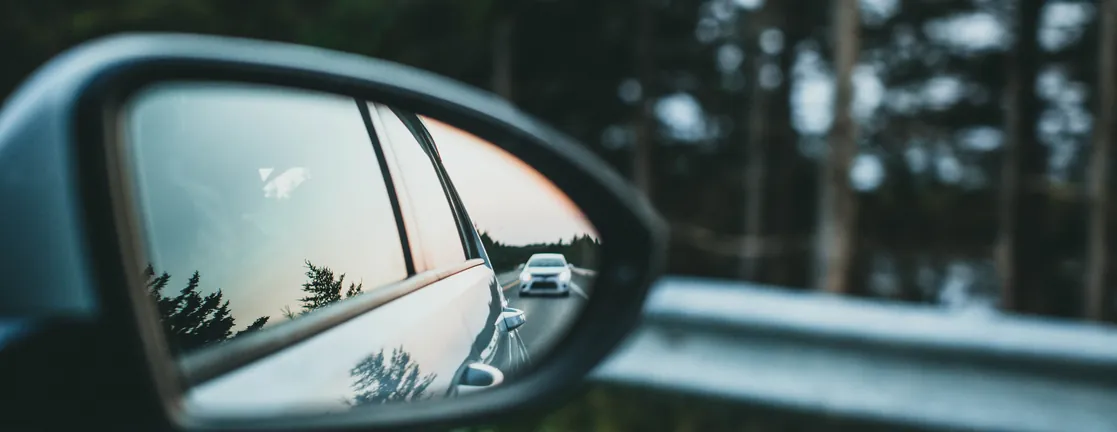 De zijspiegel van een auto, met daarin een andere auto op een weg door het bos