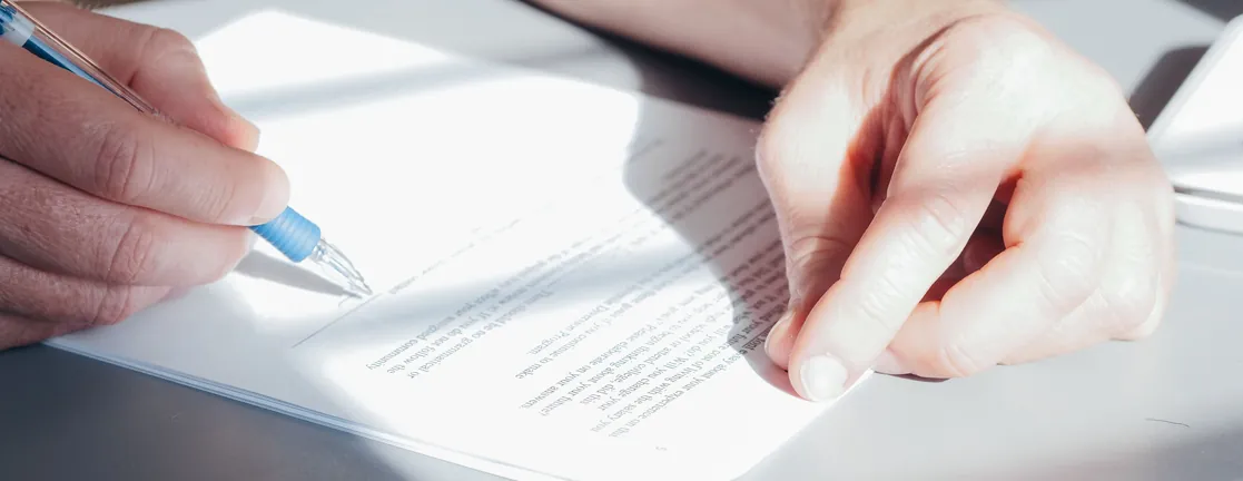 Een hand met een pen zet een handtekening onder een document op tafel