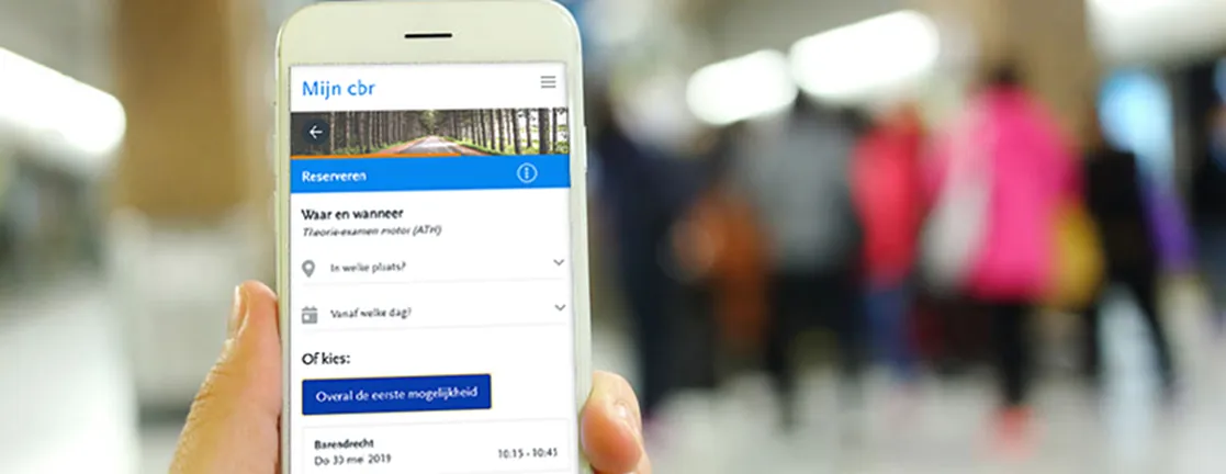 Een hand houdt een telefoon met het reserveringsscherm van Mijn CBR vast, in een drukke hal